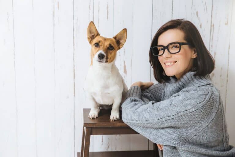 Adorable satisfied female wears spectacles and loose warm sweater, stands near her favourite dog